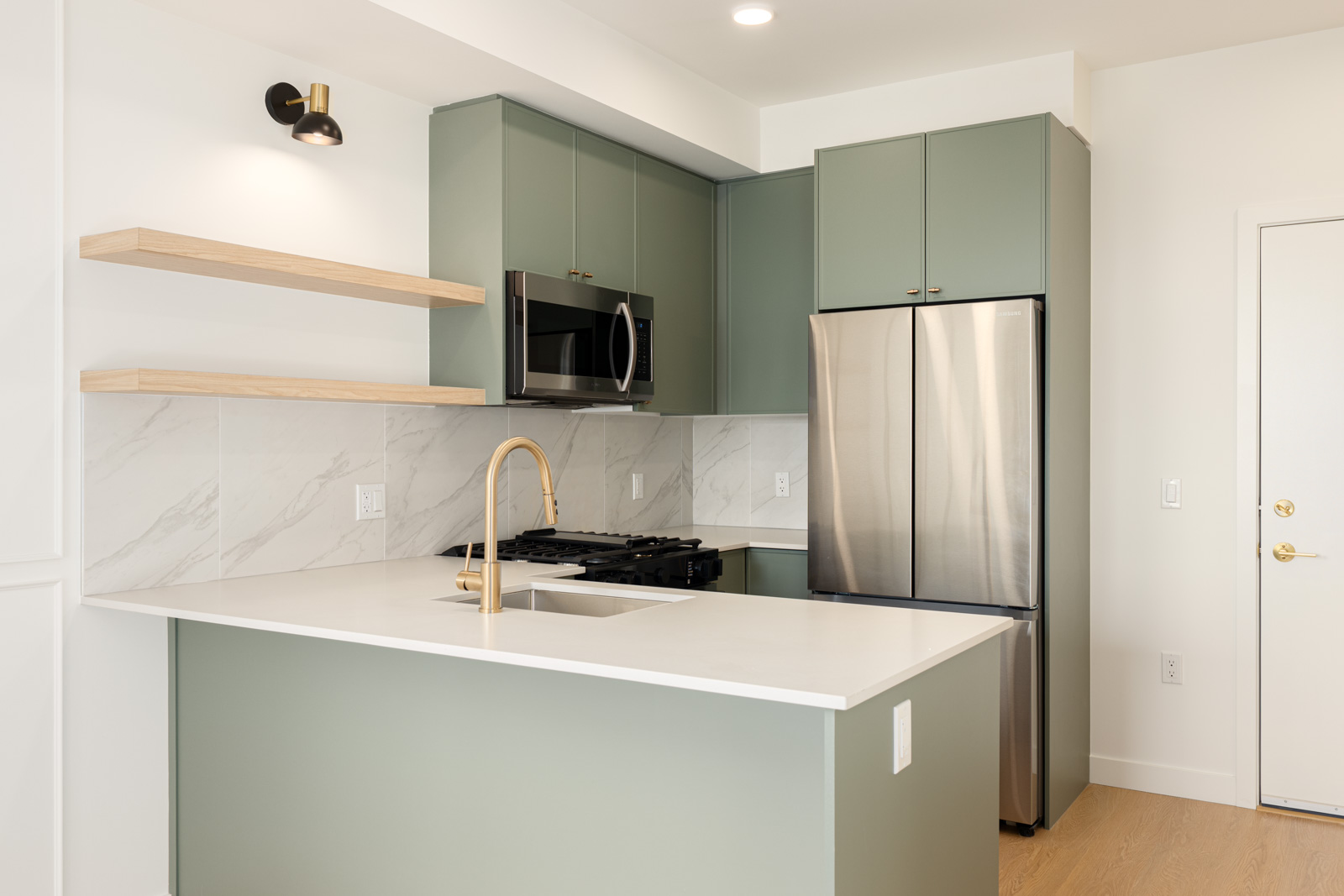 Modern kitchen with sage green cabinets, stainless steel appliances, white countertops, open wooden shelves, and a gold faucet.