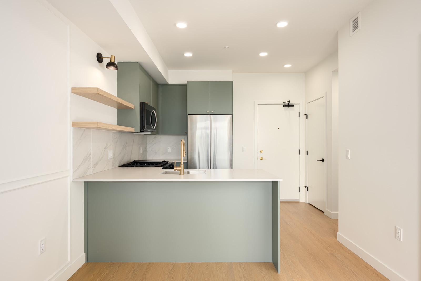 Modern kitchen with green cabinets, stainless steel appliances, open wooden shelves, a white countertop, and light wood flooring.