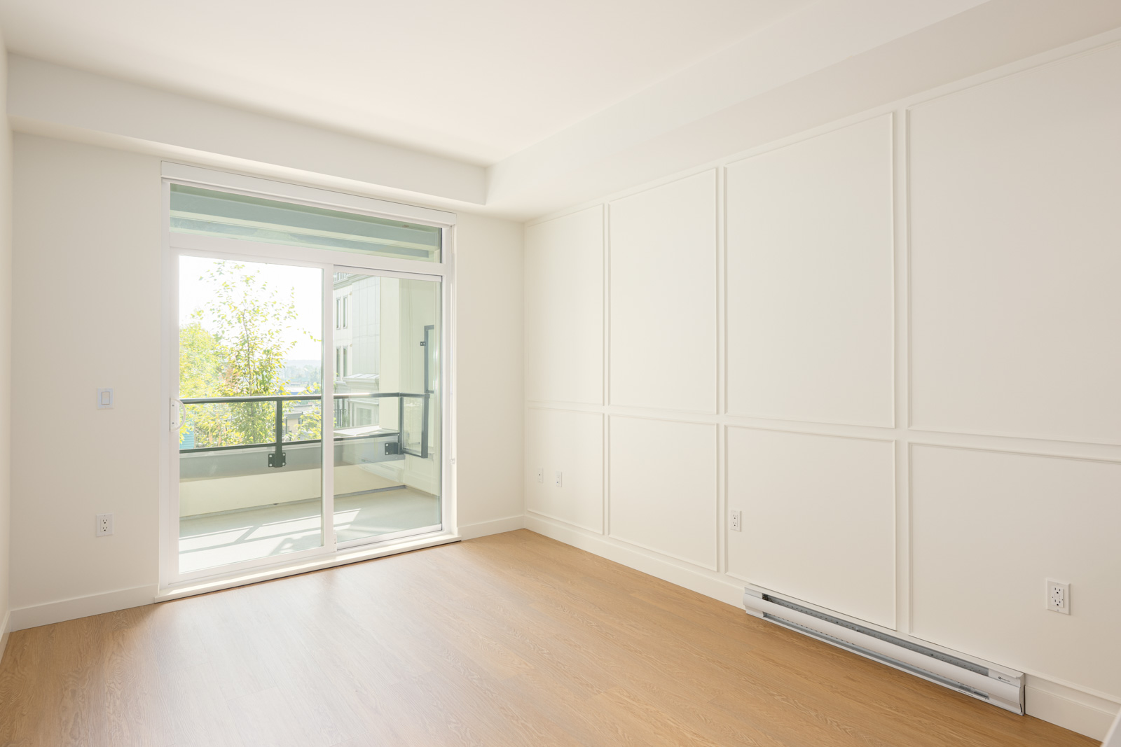 Empty room with light wood flooring, white paneled walls, and a large glass door leading to a balcony with outdoor views.