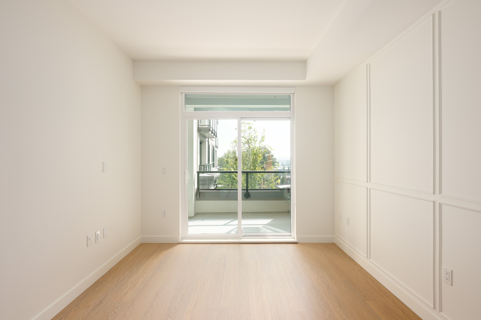 Empty room with light-colored walls and wood floor, large sliding glass door opening to a balcony with a partial view of trees and buildings outside.