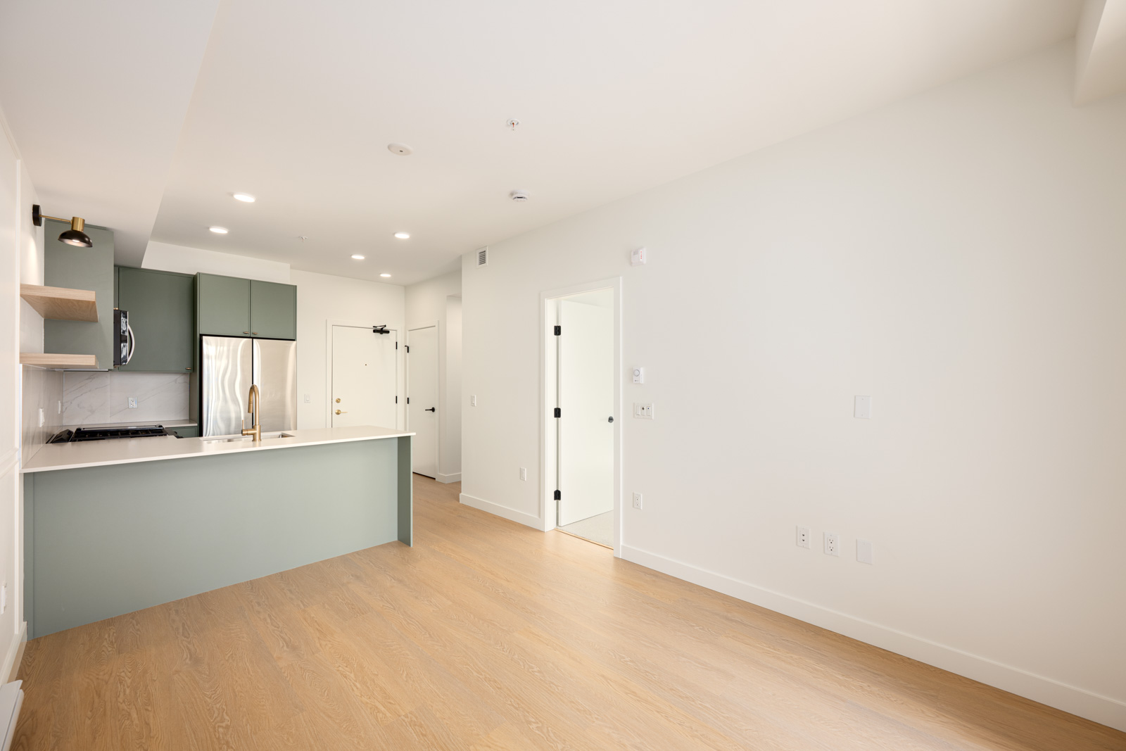 Modern apartment interior with light wood flooring, white walls, an open kitchen with green cabinets, stainless steel appliances, and two white doors in the background.