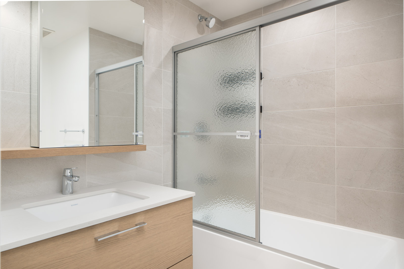 Modern bathroom with light wood vanity, white countertop, wall-mounted mirror cabinet, and bathtub with frosted glass sliding doors against light-colored tiled walls.