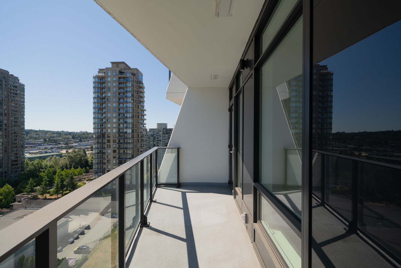 A modern apartment balcony with glass railing, overlooking city buildings and a green area under a clear blue sky during daytime.