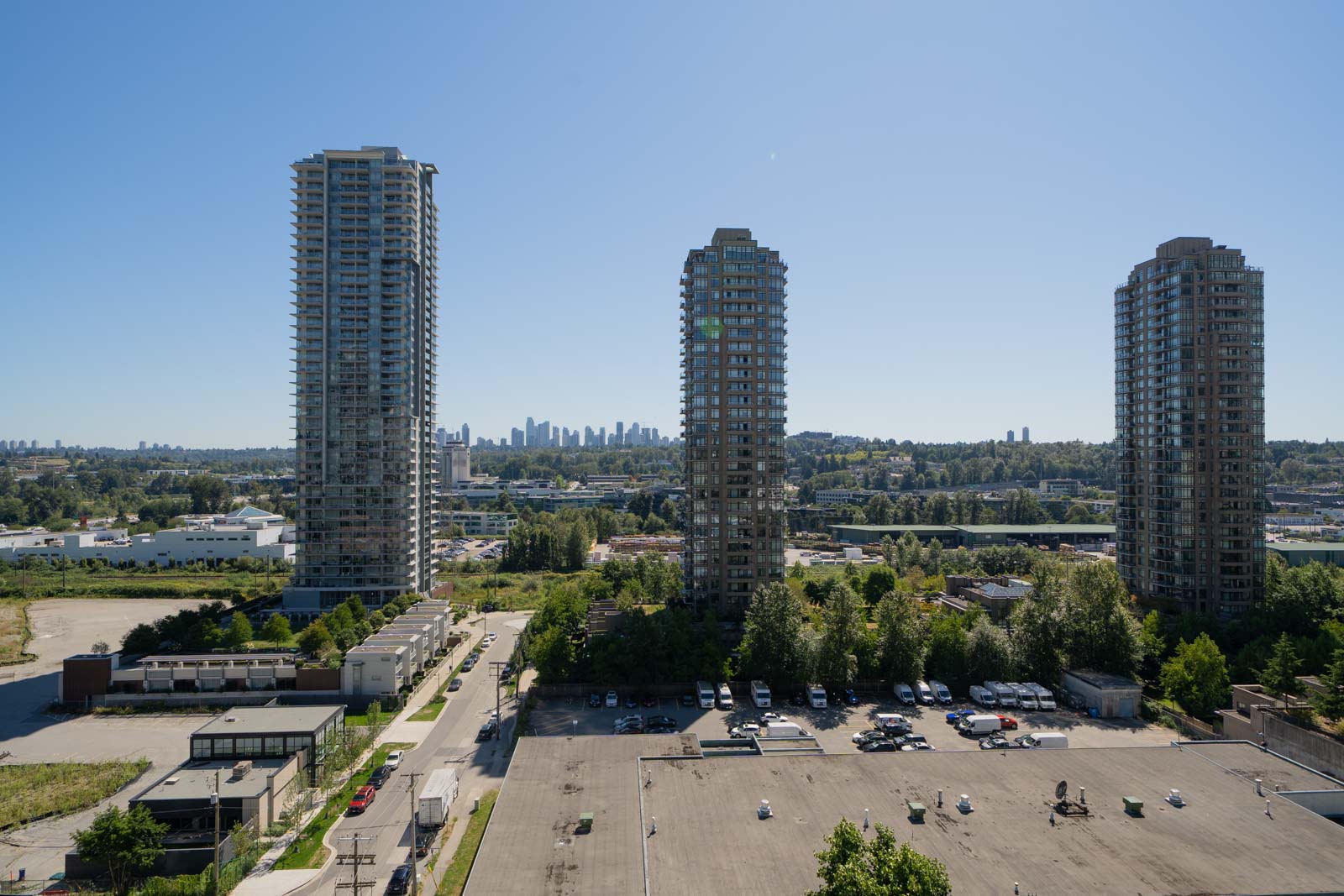 Three tall residential buildings stand near a parking lot, with trees and a city skyline visible in the background under a clear blue sky.