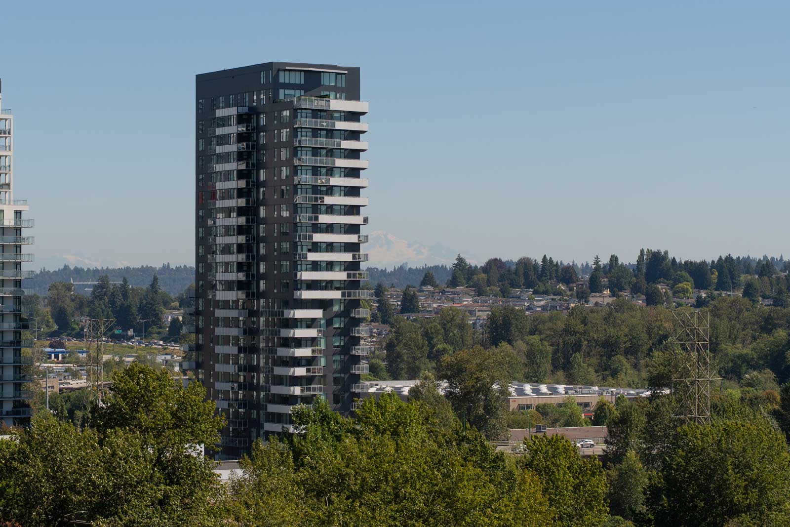A tall modern apartment building stands among trees and low-rise structures, with a distant mountain barely visible in the background under a clear sky.
