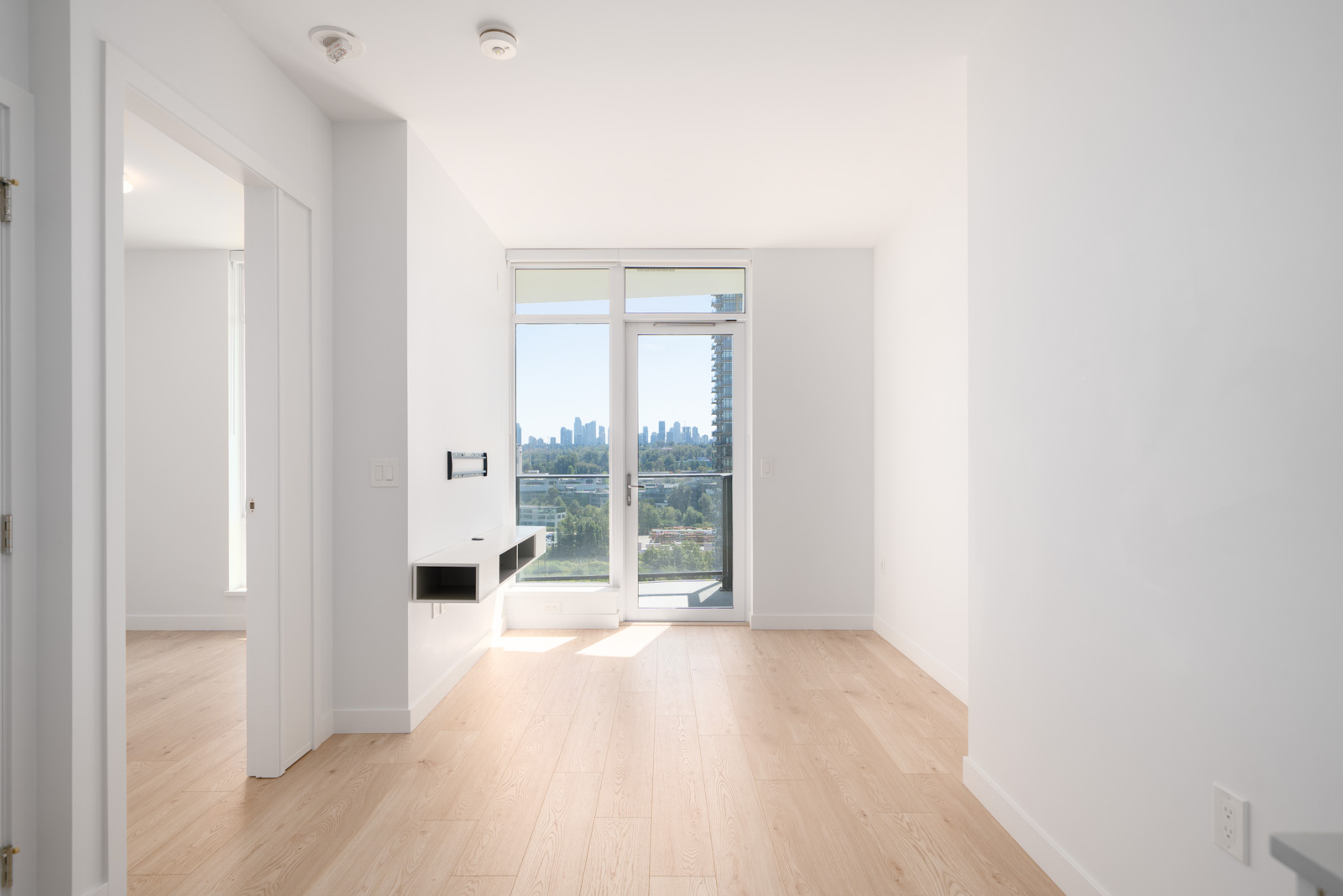 Bright, empty room with light wood flooring, white walls, a built-in shelf, large window, and glass door leading to a balcony with a city skyline view.