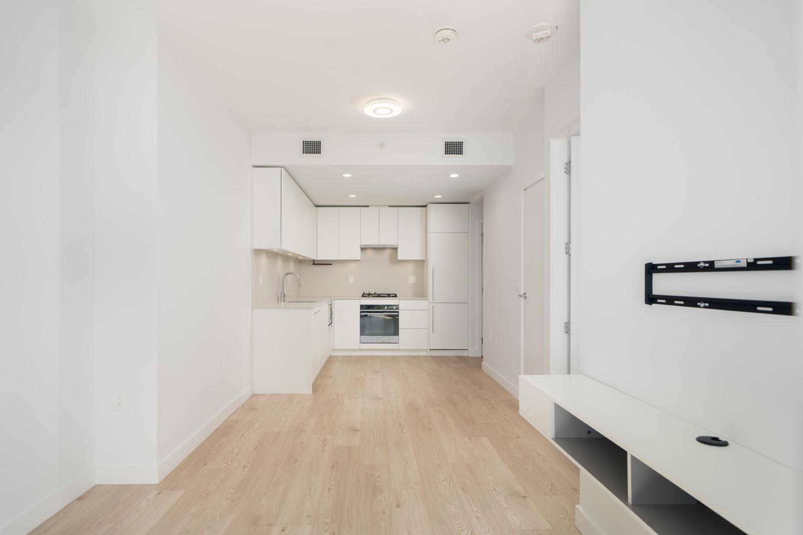 Minimalist kitchen with white cabinets, light wood flooring, and built-in oven, located in an open-plan apartment with neutral walls and mounted shelves on the right.