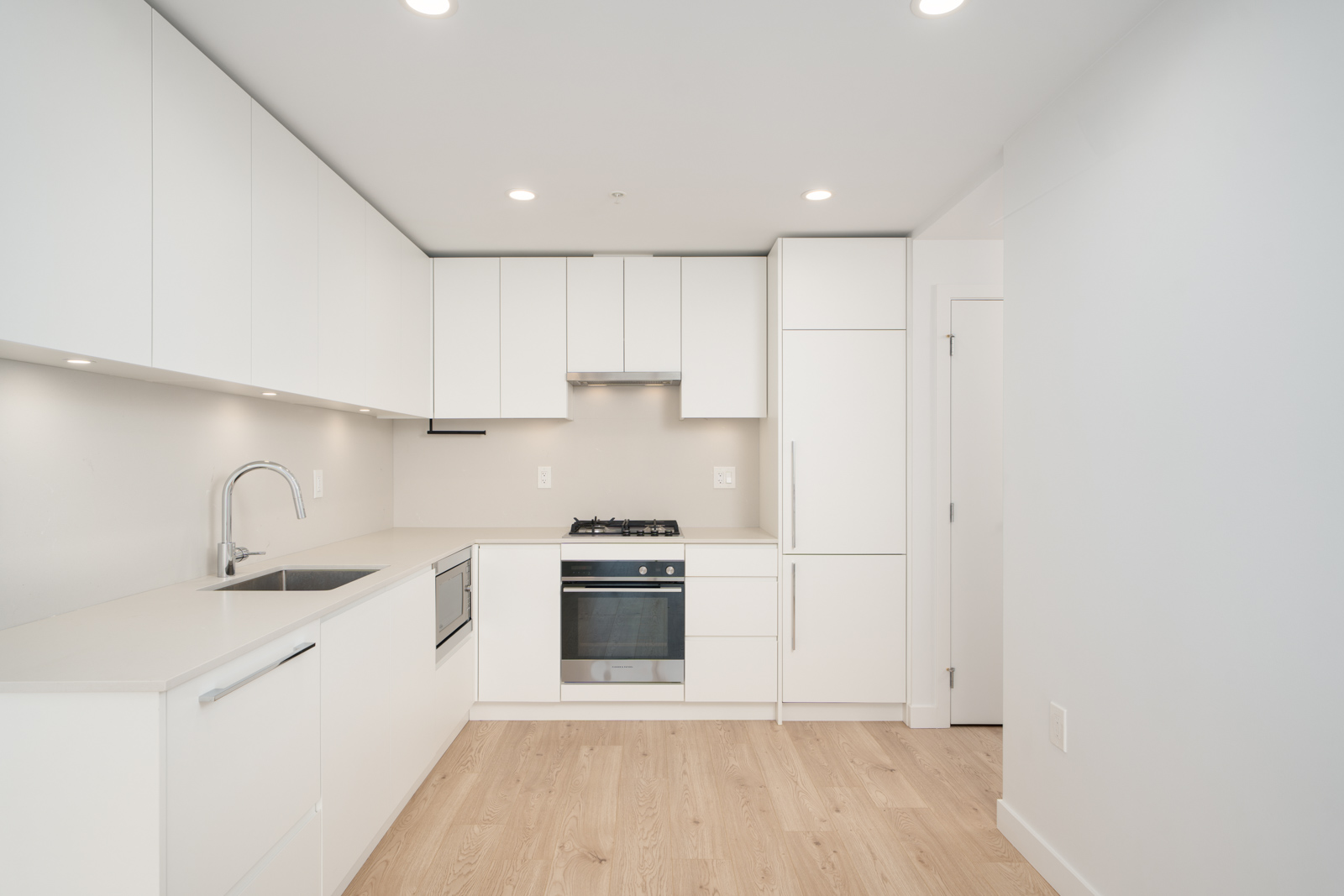 Modern kitchen with white cabinets, built-in oven, gas cooktop, stainless steel sink, and light wood flooring under recessed ceiling lights.