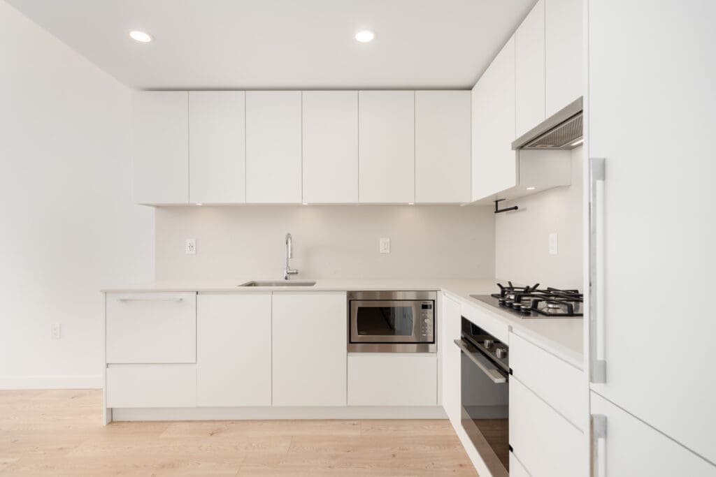 A modern, minimalist kitchen with white cabinets, integrated appliances, a gas stove, microwave, and light wood flooring.
