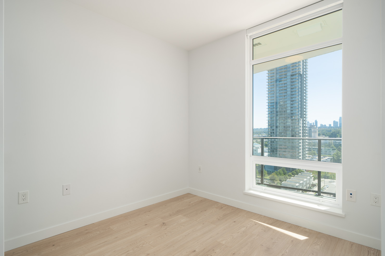 Empty, unfurnished room with light-colored wooden floor, white walls, and a large window overlooking tall buildings and a cityscape on a clear day.