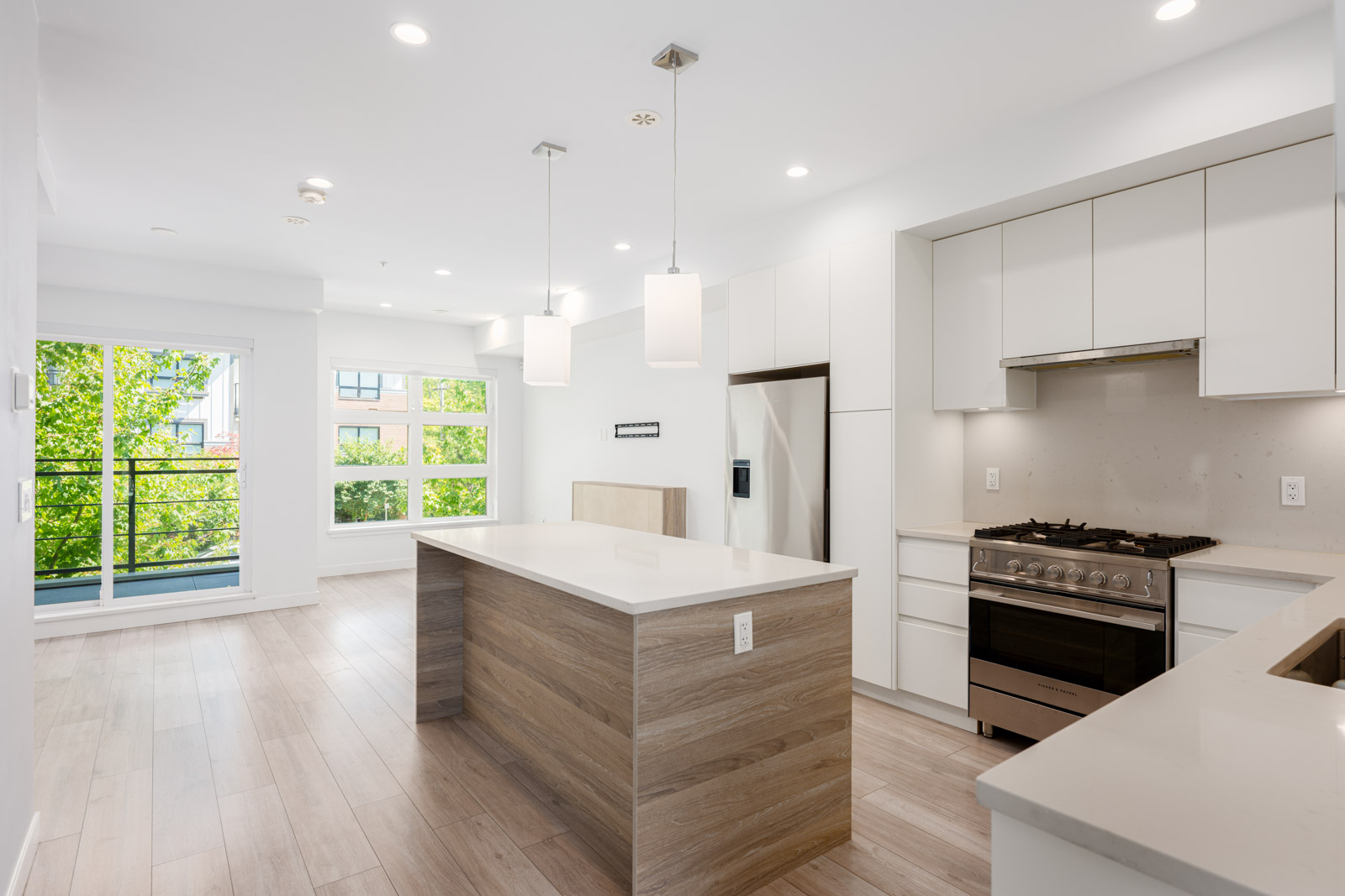 Modern, unfurnished kitchen with white cabinets, stainless steel appliances, an island with wood finish, pendant lights, and large windows overlooking greenery.