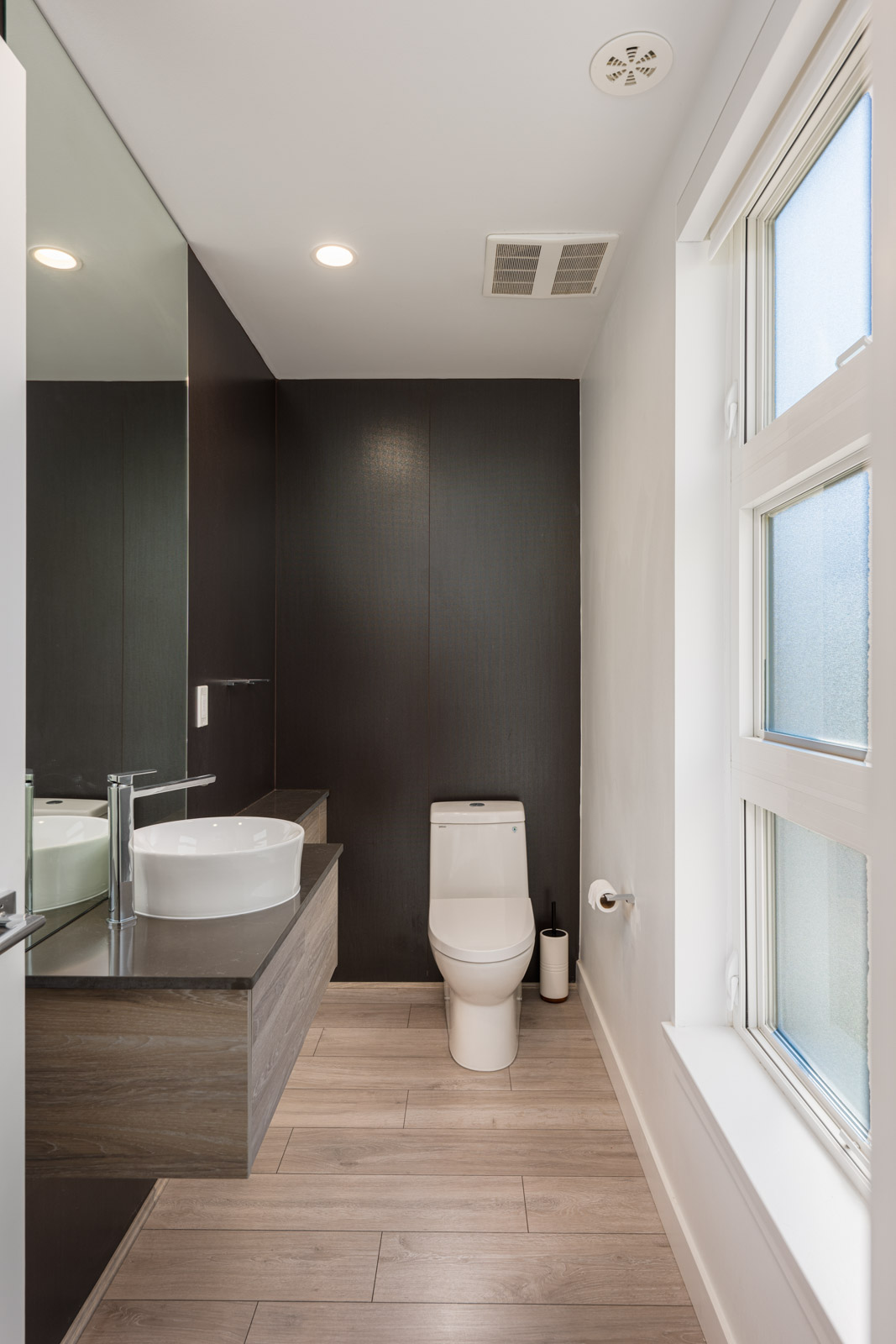 Modern bathroom with a floating vanity, vessel sink, wall mirror, toilet, and large frosted window; light wood flooring and dark accent wall.