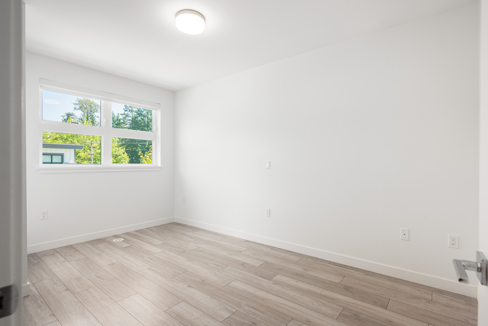 Empty room with white walls, wood-look tile flooring, a ceiling light, and three rectangular windows showing greenery outside.