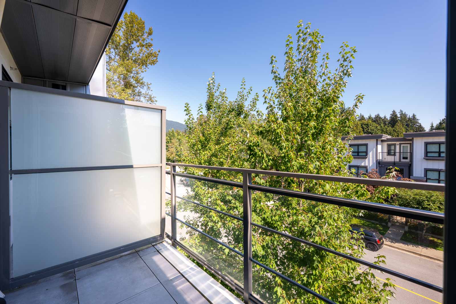 A small balcony with metal railing and frosted glass divider overlooks green trees and a quiet residential street on a clear, sunny day.
