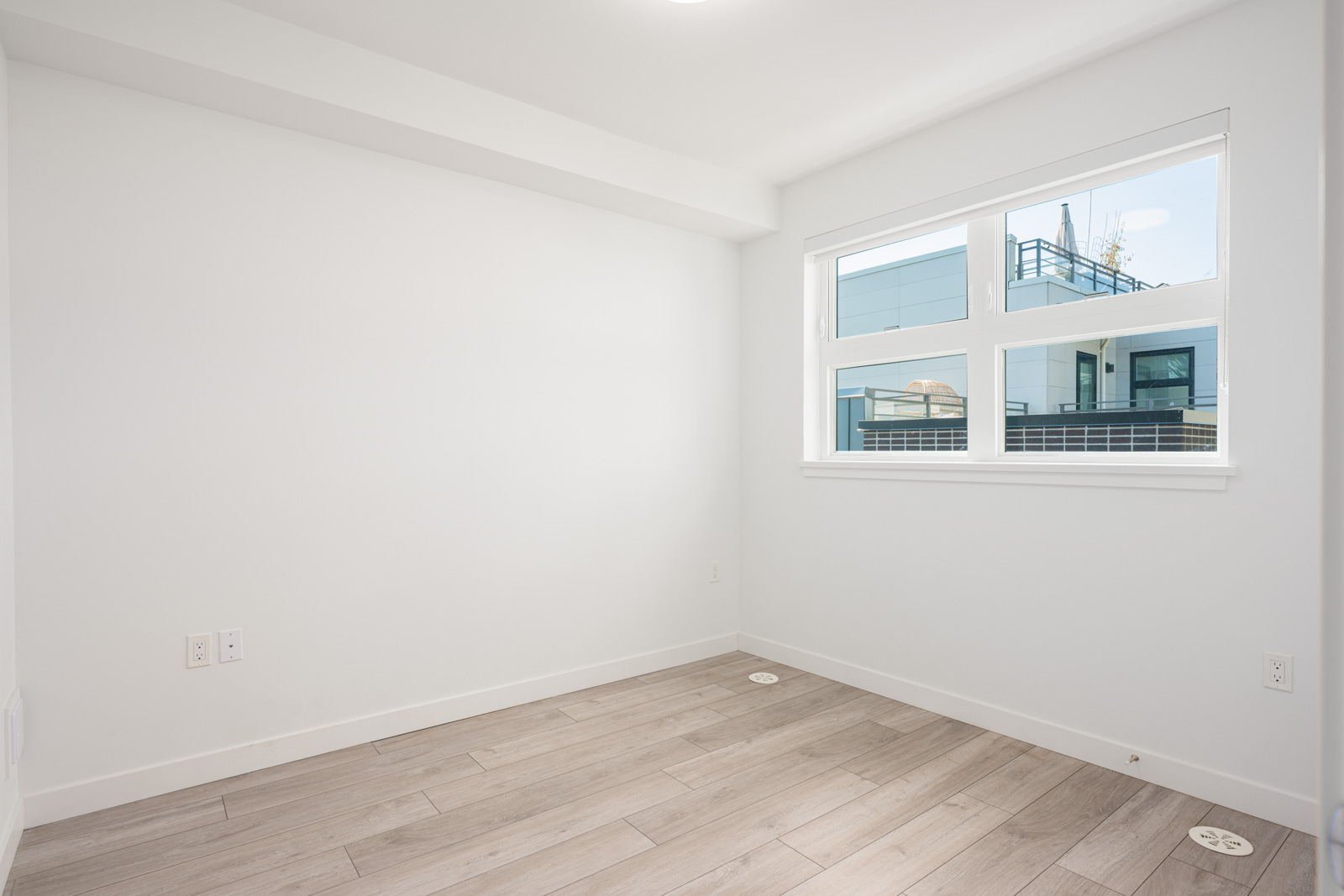 Empty room with light wood flooring, white walls, and a window showing an outside modern building. Electrical outlets are visible on the walls.