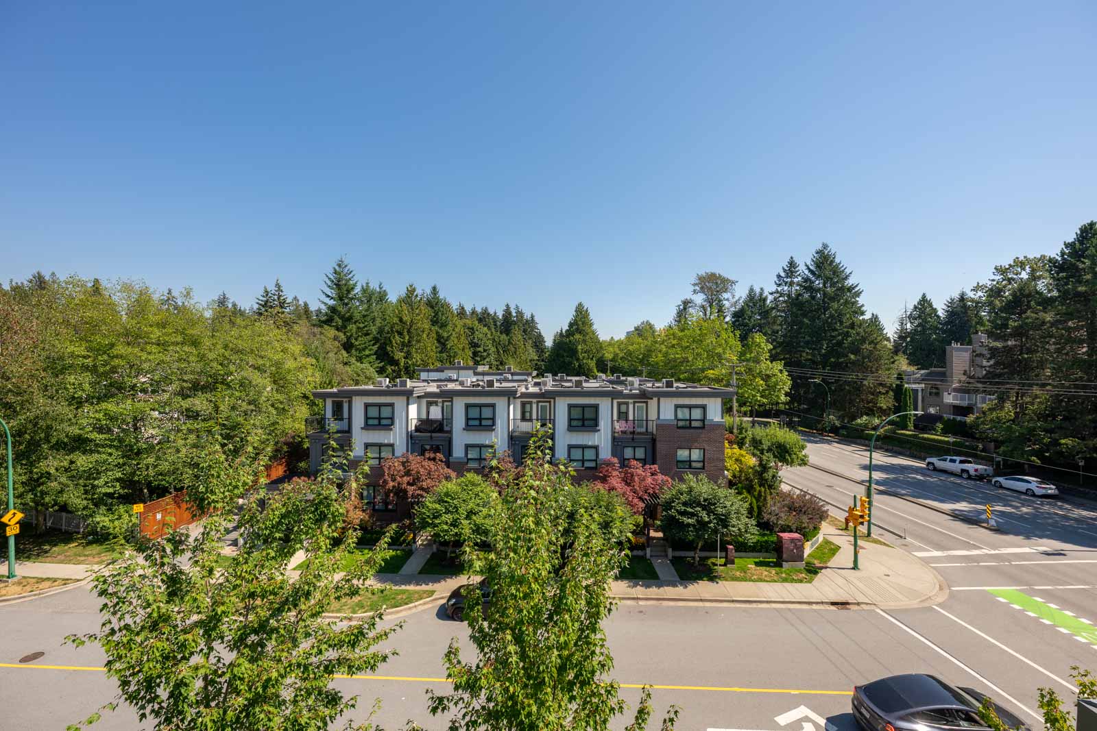Modern three-story apartment building surrounded by trees at a street intersection on a clear, sunny day. Cars are visible on the road.