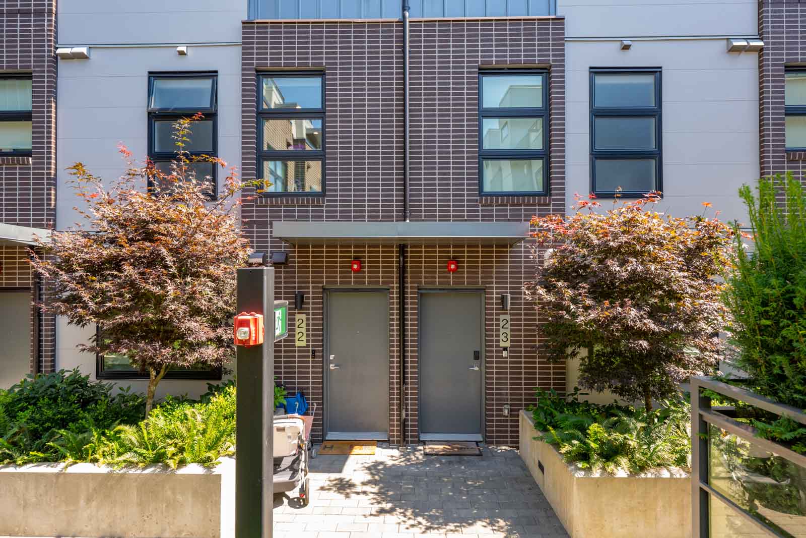 Front view of two modern townhouse entrances, units 122 and 123, with brick facade, grey doors, small trees, and planters on a sunny day.