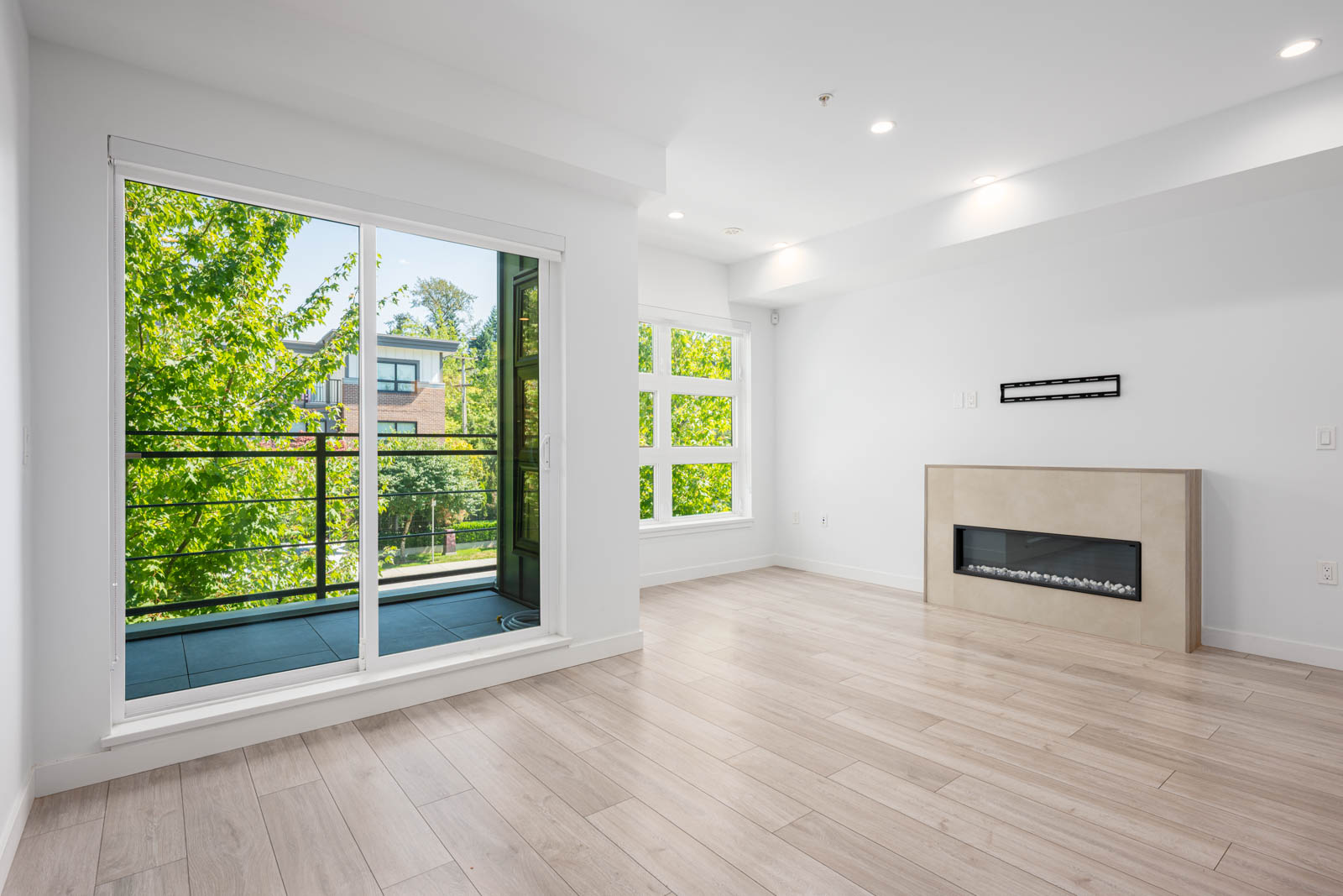 Bright, empty living room with light wood flooring, modern fireplace, large windows, and sliding glass door leading to a balcony with green trees outside.