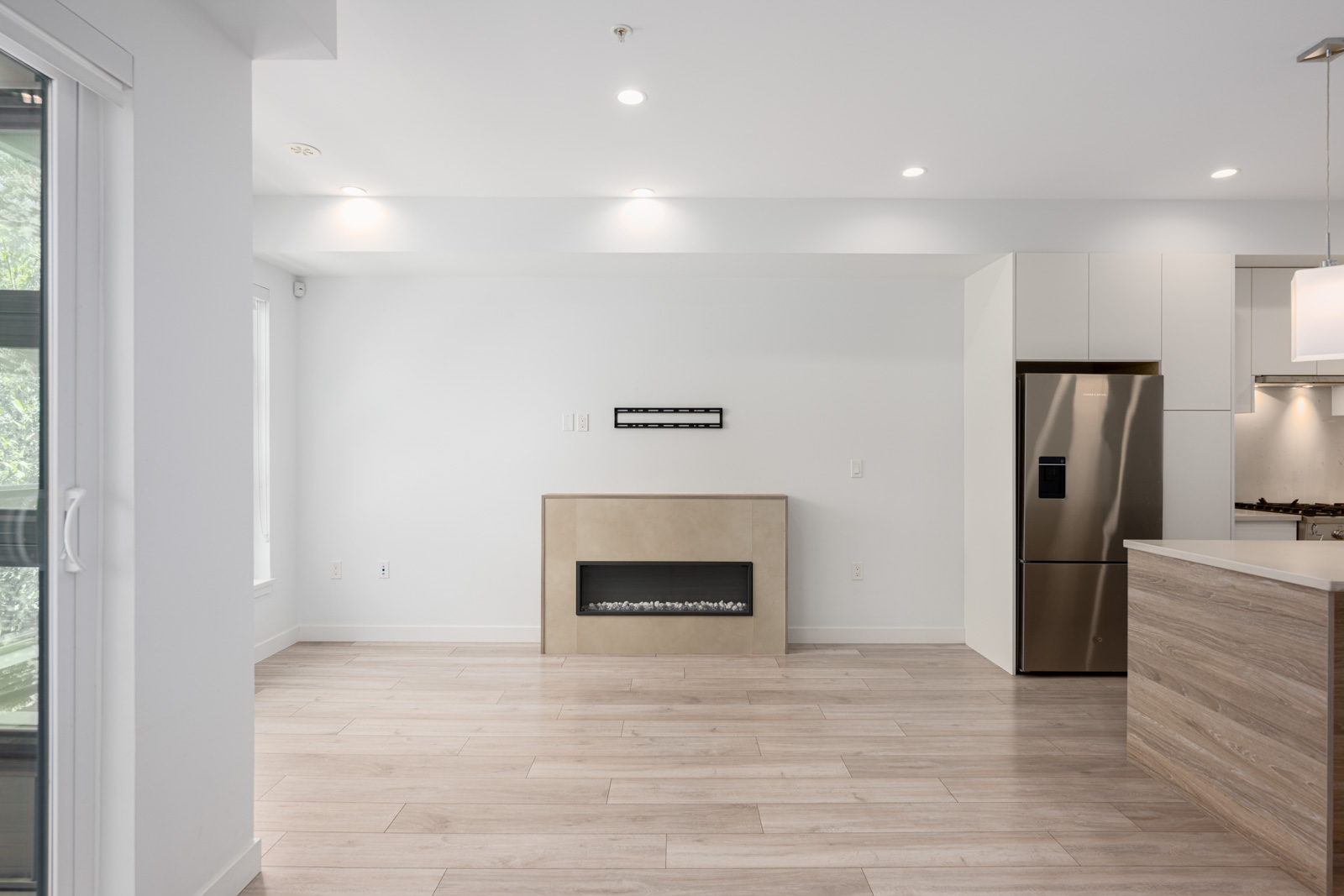 Modern, unfurnished living space with light wood floors, a wall-mounted electric fireplace, recessed lighting, and a view of the kitchen with stainless steel appliances.
