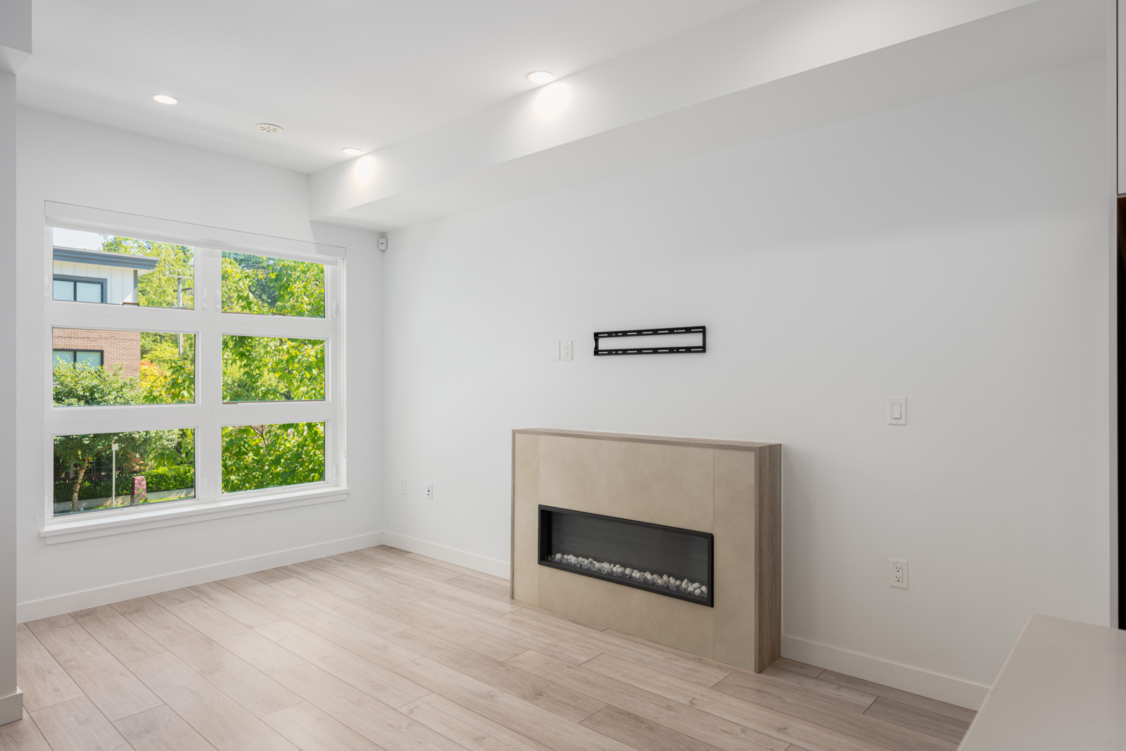 Bright, modern living room with large window, light wood flooring, recessed lighting, and a minimalist fireplace below a mounted bracket on a white wall.