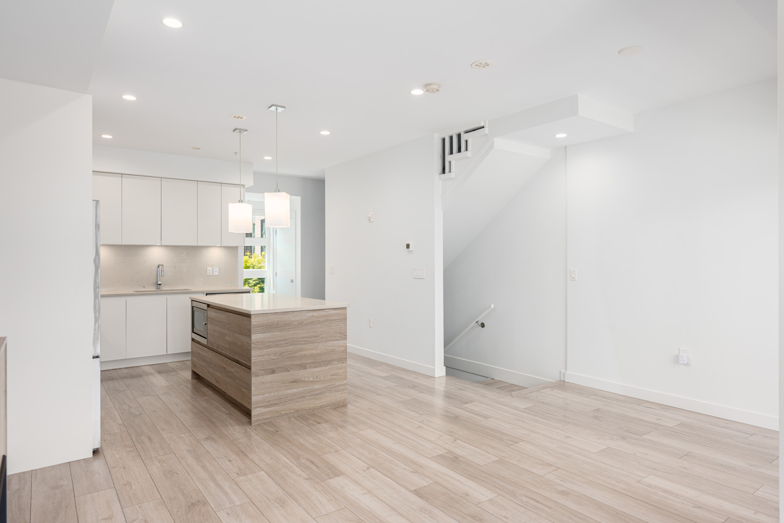 Modern kitchen with light wood floors, a central island, white cabinets, and pendant lights, adjacent to an open stairway and entryway in a bright, minimalist interior.