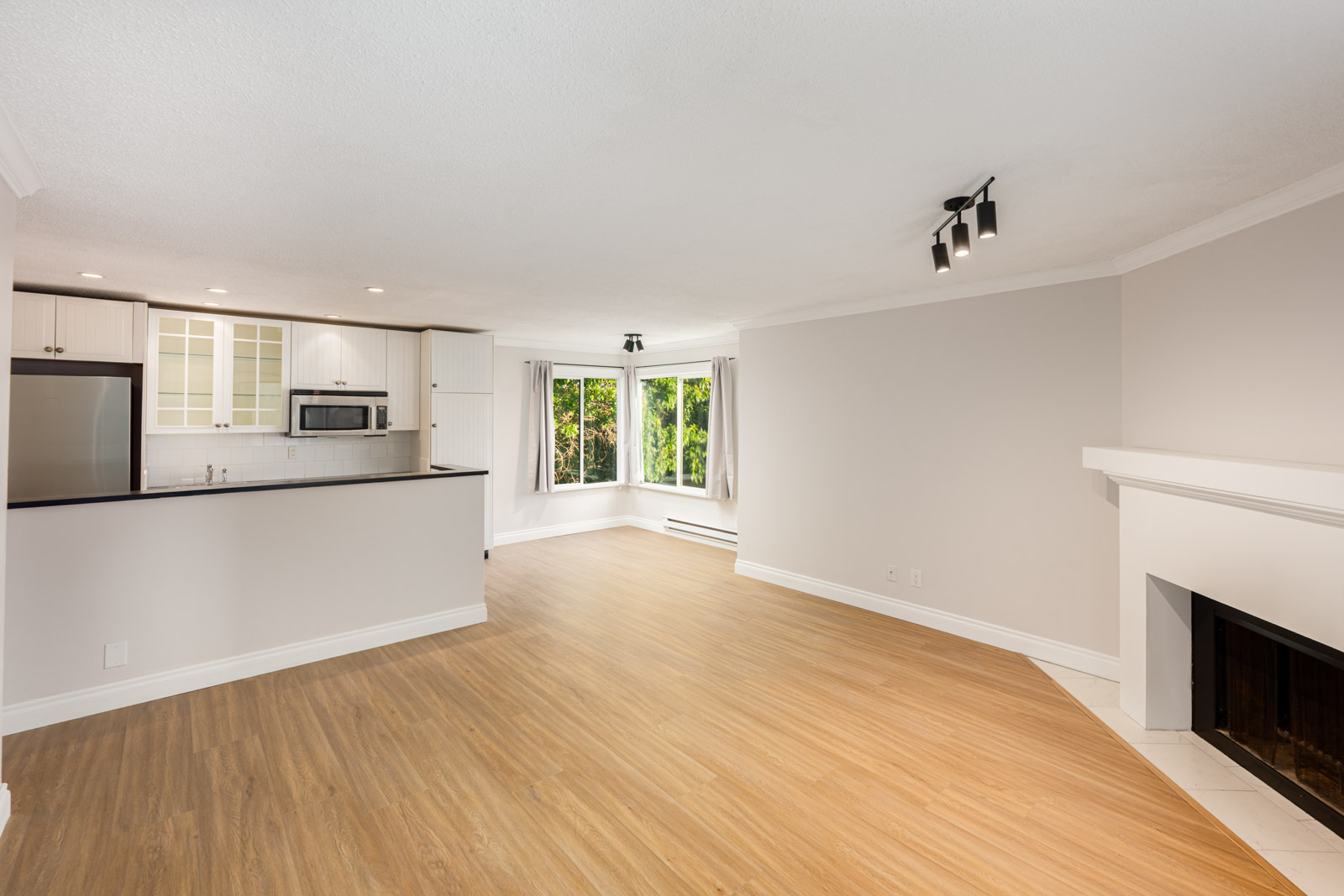 Open-concept living area with light wood floors, white walls, kitchen with stainless steel appliances, bay window, and fireplace.