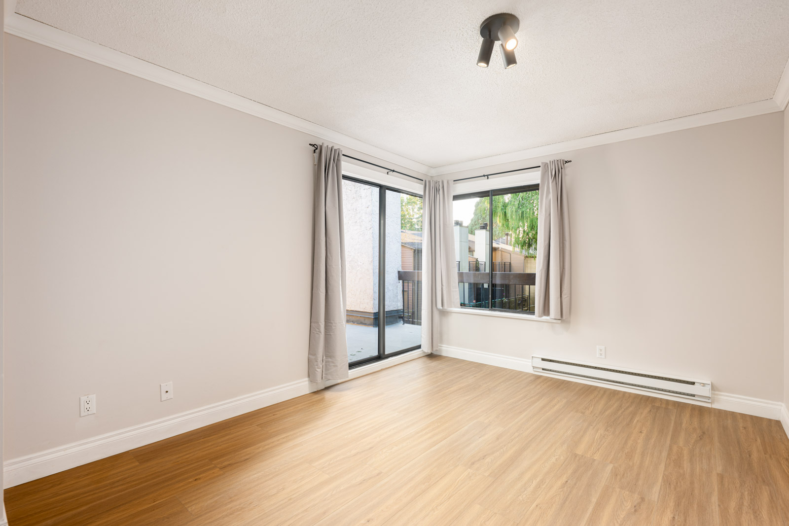 Empty room with light wood flooring, white walls, ceiling light fixture, wall heater, and large corner windows with gray curtains overlooking an outdoor space.