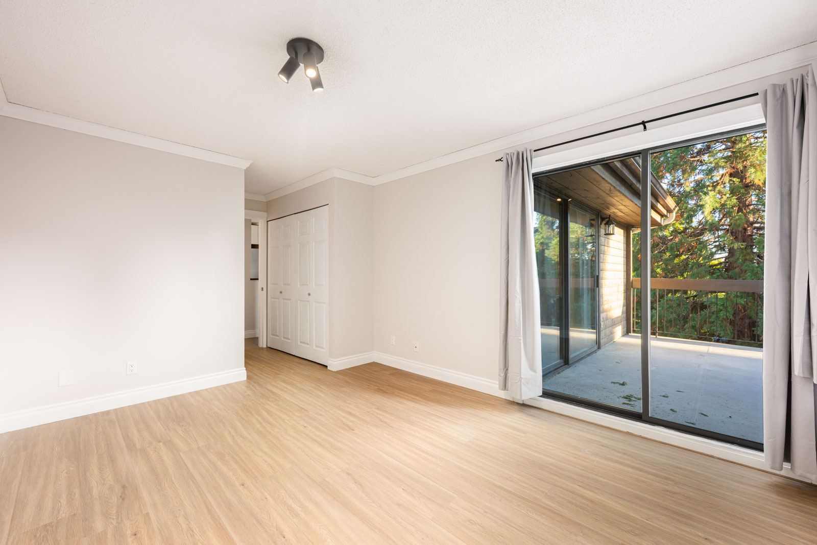 Empty room with light wood flooring, grey walls, a closet with white doors, and a large sliding glass door opening to a balcony with trees visible outside.