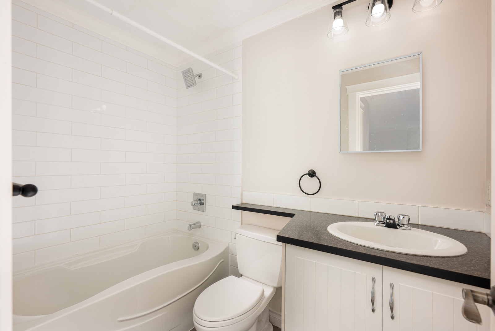 A small bathroom with a bathtub, toilet, and sink with black countertop, a wall mirror, and white tiled walls. Three light fixtures are above the mirror.