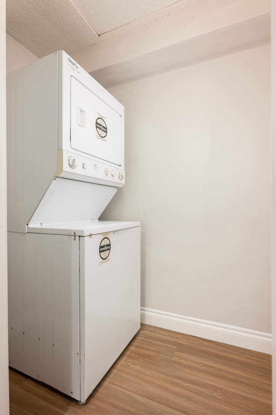 A white stacked washer and dryer unit is positioned against a beige wall on a wood floor in a small laundry area.