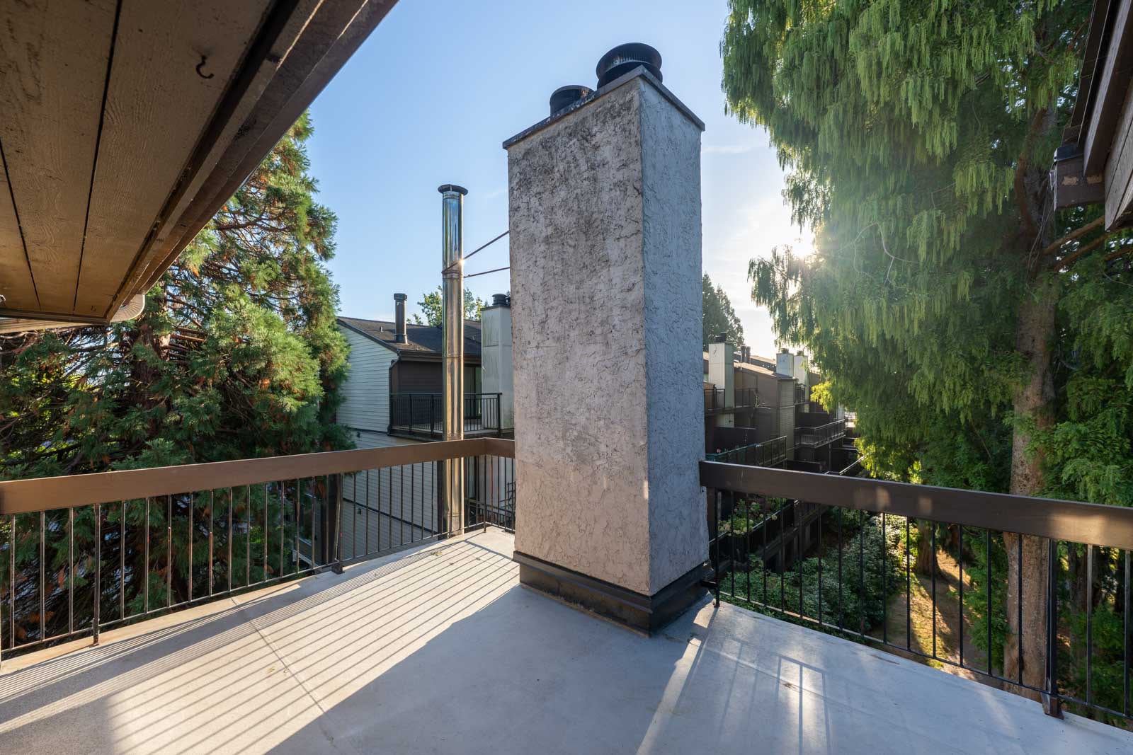 A concrete balcony with metal railings features a large chimney in the center, surrounded by trees and adjacent residential buildings in daylight.
