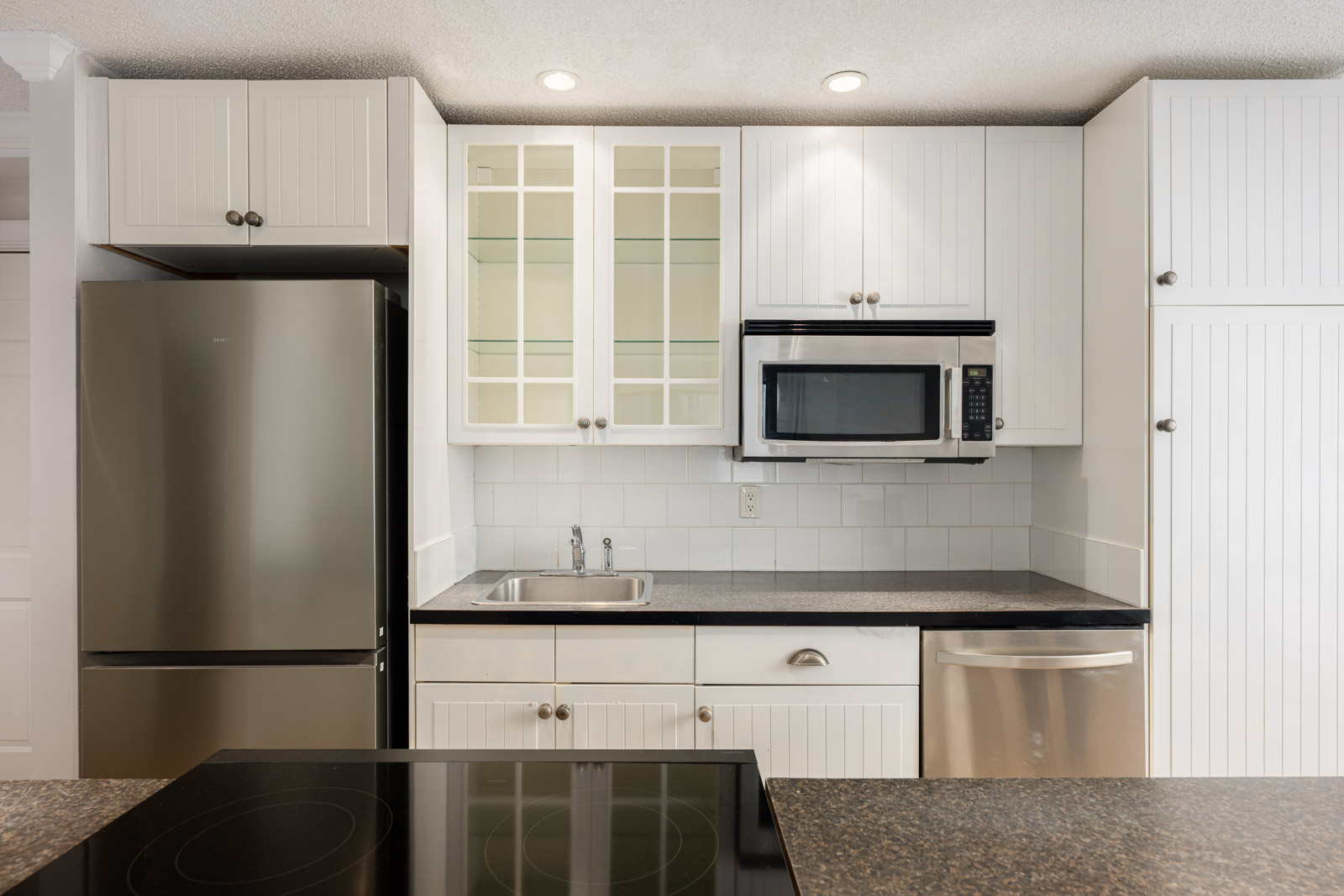 Modern kitchen with white cabinets, stainless steel refrigerator and dishwasher, microwave above sink, glass-front cabinets, and electric stove in foreground.