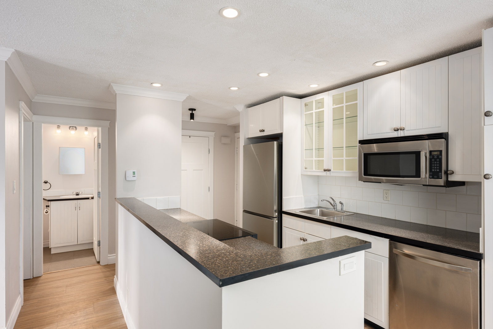 Modern kitchen with white cabinets, stainless steel appliances, black countertops, and a breakfast bar; hallway leads to another room with a vanity and mirror.