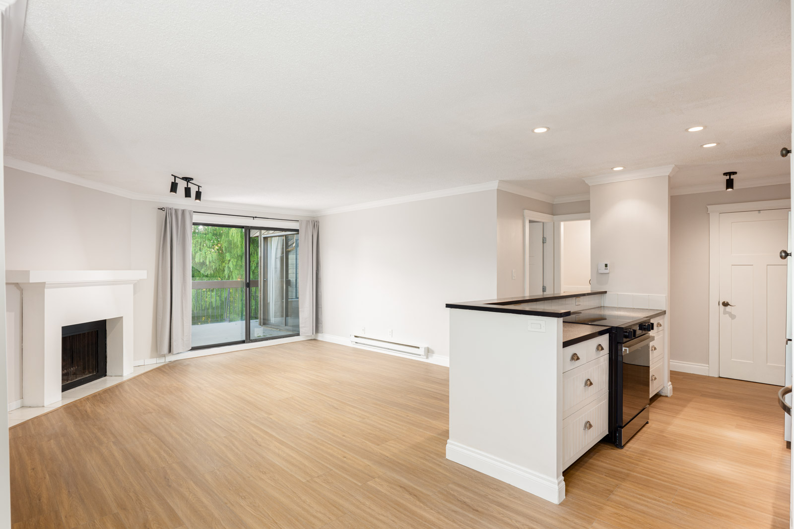 Open-plan living area with light wood flooring, white walls, a fireplace, large windows with balcony access, and a modern kitchen with black countertops and white cabinetry.