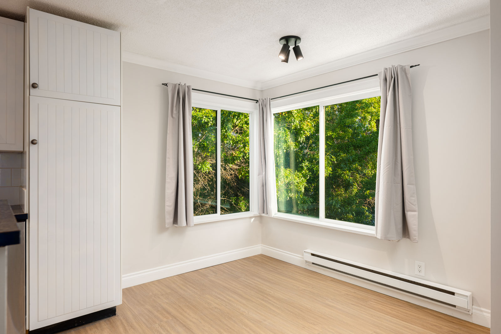Bright, empty corner of a room with large windows, light gray curtains, wood flooring, white walls, and a ceiling light fixture. Lush green trees are visible outside.