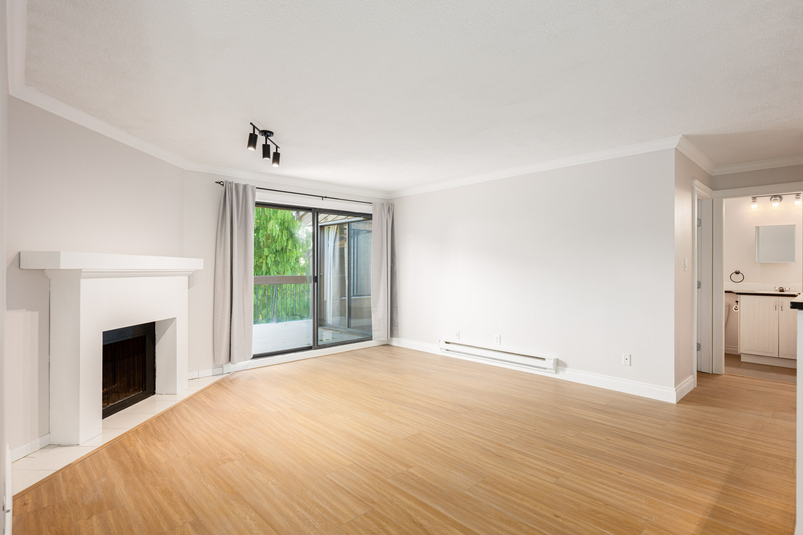 Unfurnished living room with light wood flooring, a white fireplace, large window with curtains, and doorway leading to a bathroom and kitchen area.