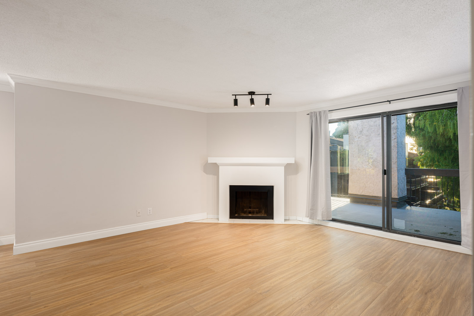 Empty living room with light wood flooring, a white fireplace, and large sliding glass doors with curtains leading to an outdoor balcony.