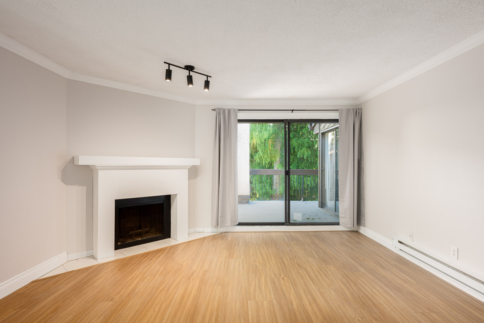 Unfurnished living room with a fireplace, wood flooring, white walls, ceiling light, and sliding glass doors leading to a balcony with greenery outside.