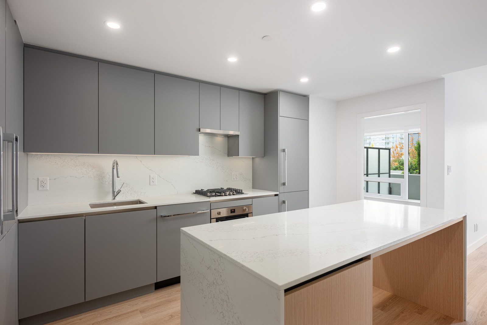 Modern kitchen with gray cabinets, built-in appliances, a marble-patterned island, under-cabinet lighting, and a large window letting in natural light.