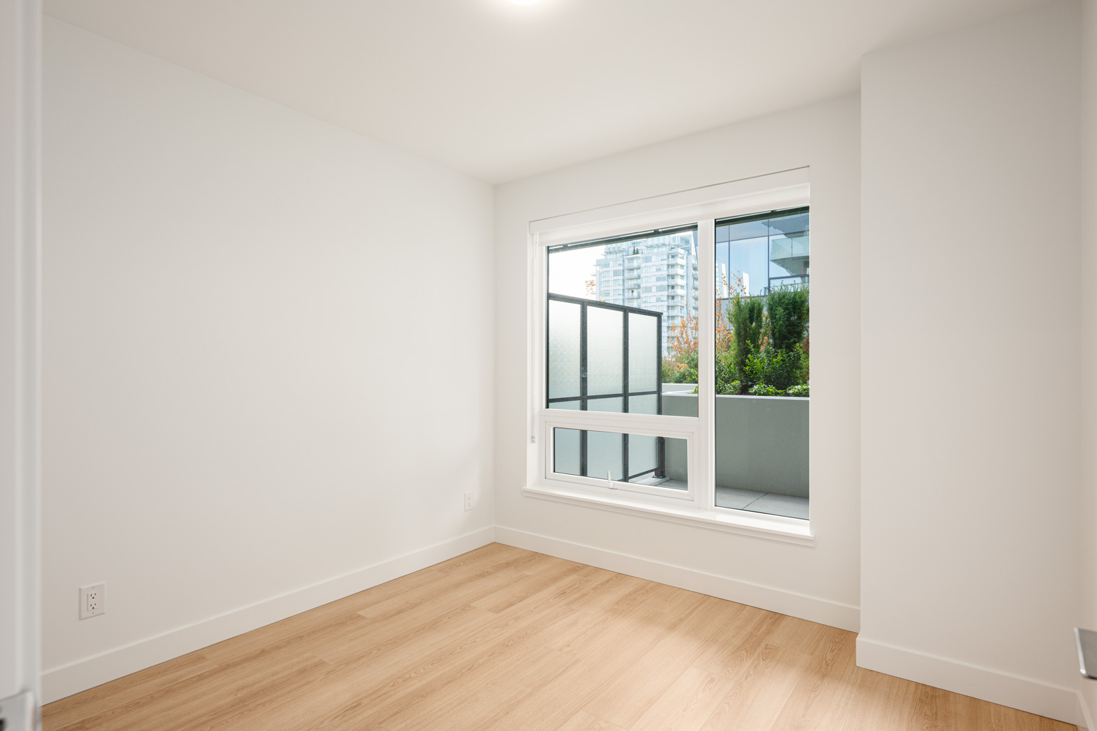 Unfurnished, empty room with light wood flooring, white walls, and a large window overlooking a balcony and city buildings.