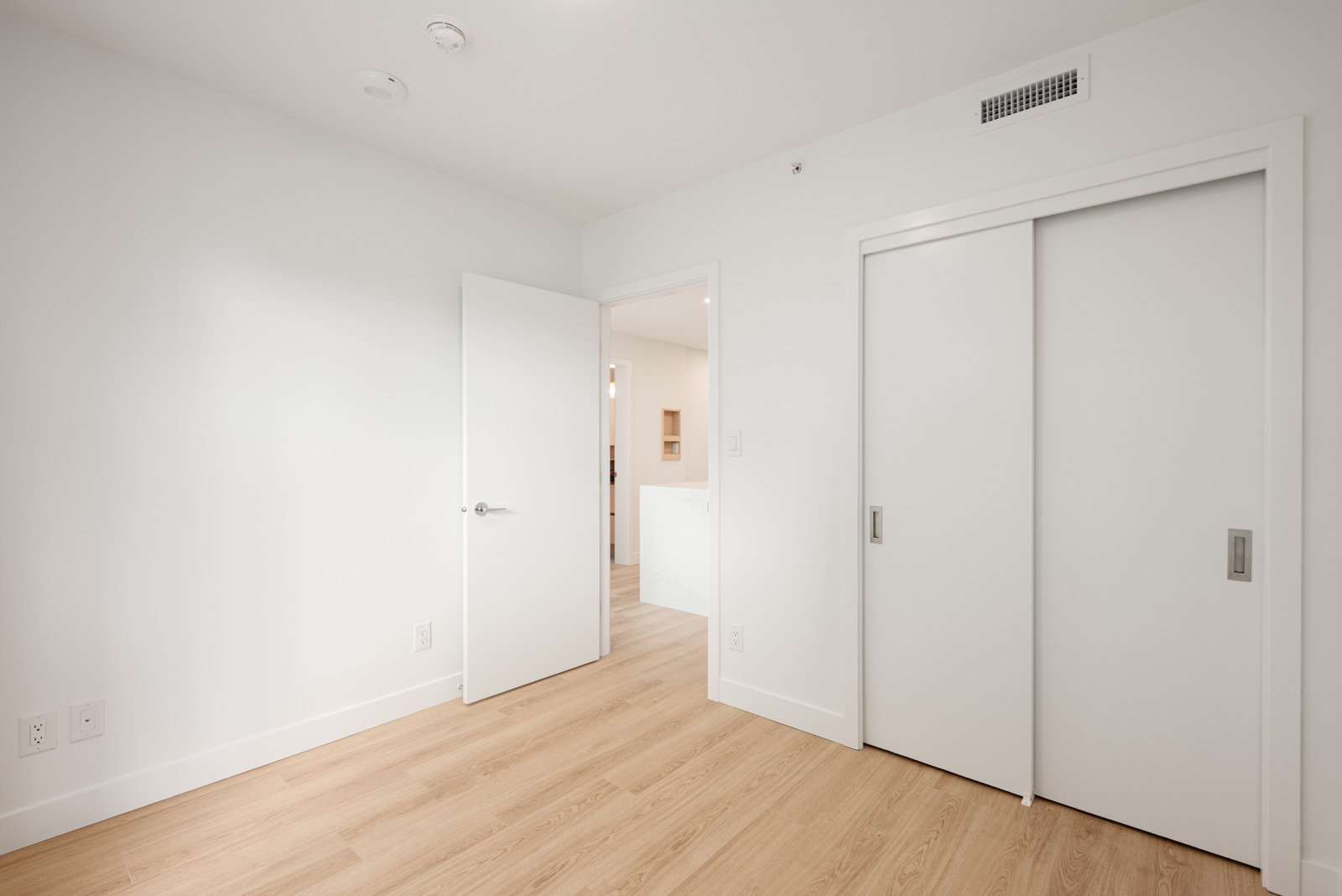 Minimalist empty room with light wood flooring, white walls, a partially open door, and a closet with sliding white doors.