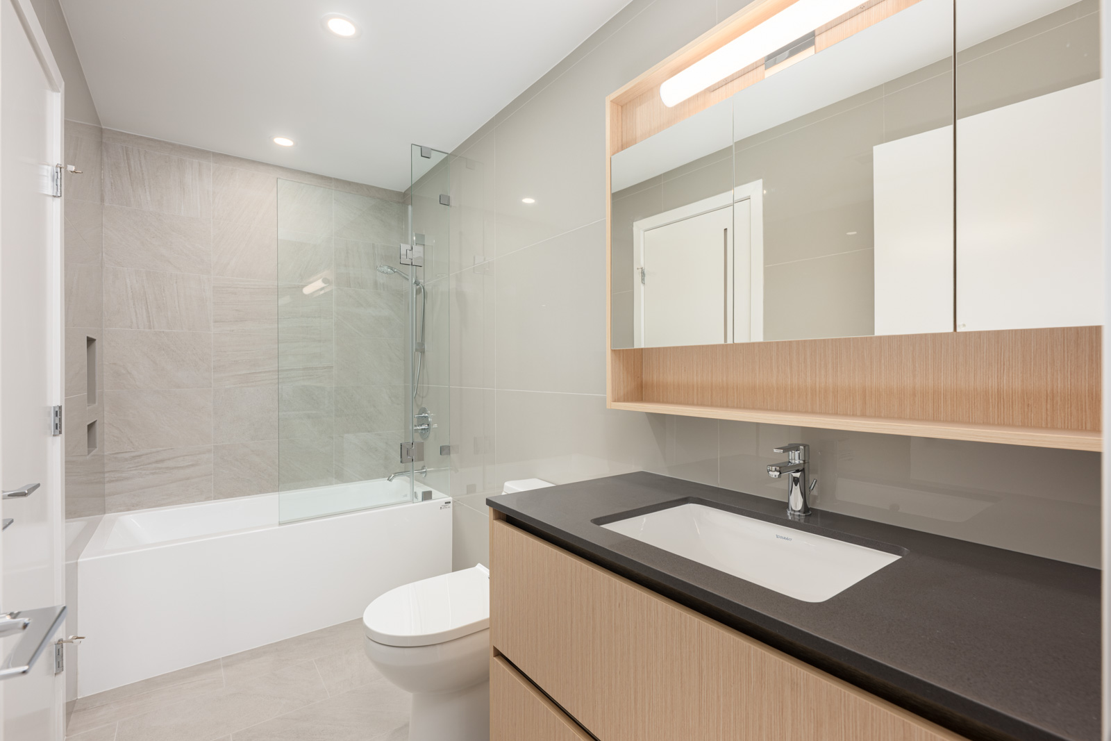 Modern bathroom with a glass-enclosed bathtub, wall-mounted vanity with sink, large mirror, wooden cabinetry, and neutral-colored tiles.