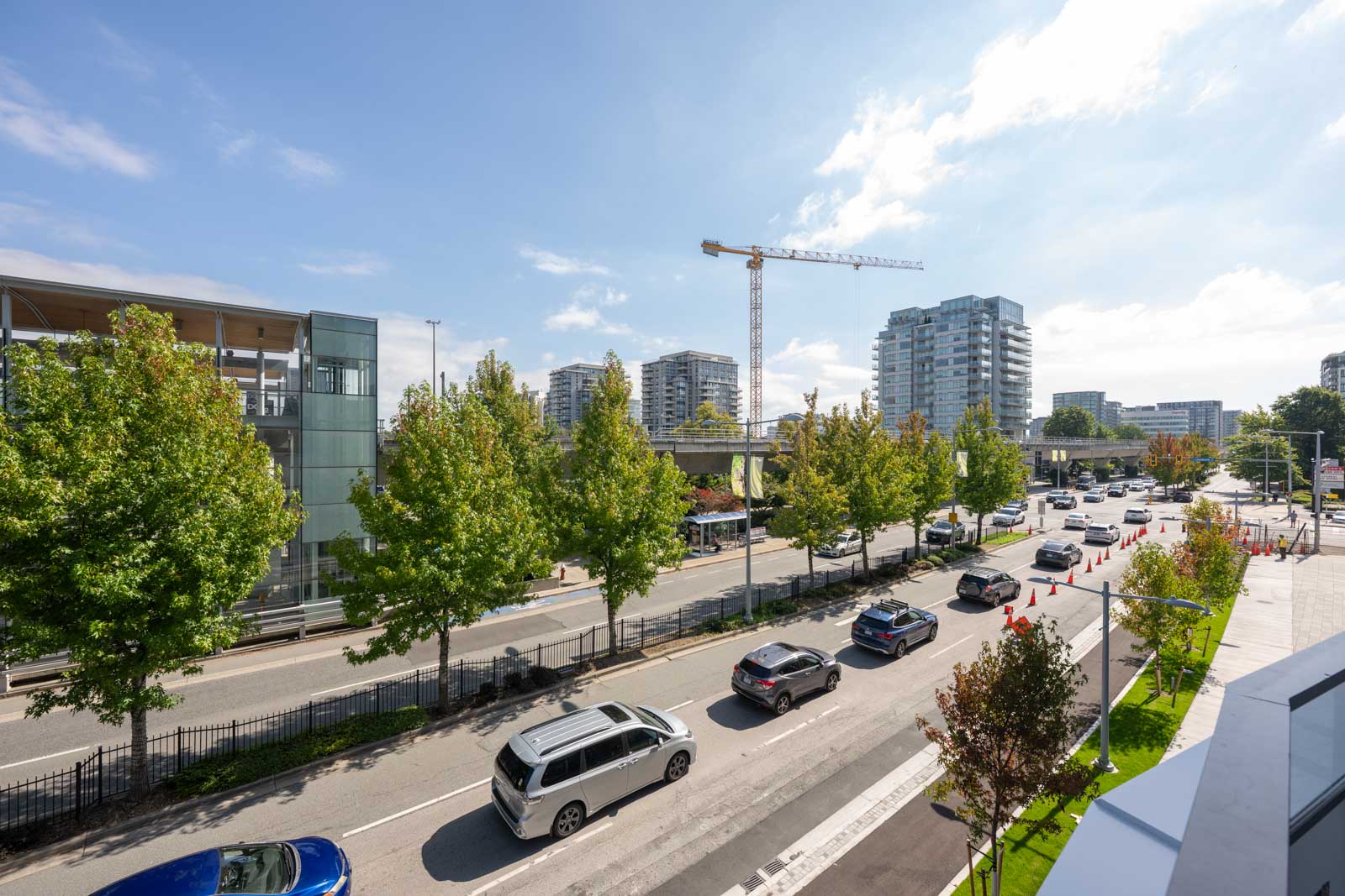 Multiple cars drive along a city street lined with trees; modern buildings and a construction crane are visible in the background under a partly cloudy sky.