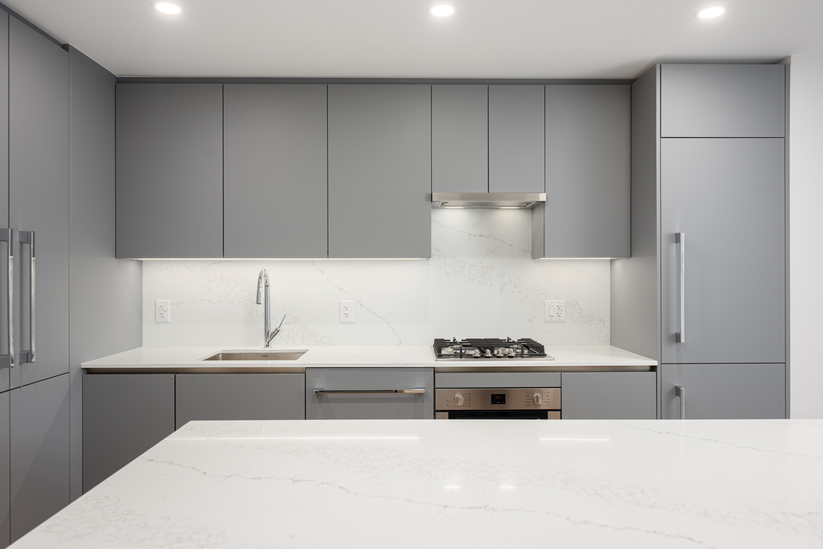 Modern kitchen with matte gray cabinets, built-in appliances, white countertops, gas stove, and under-cabinet lighting. The overall design is minimalist and uncluttered.