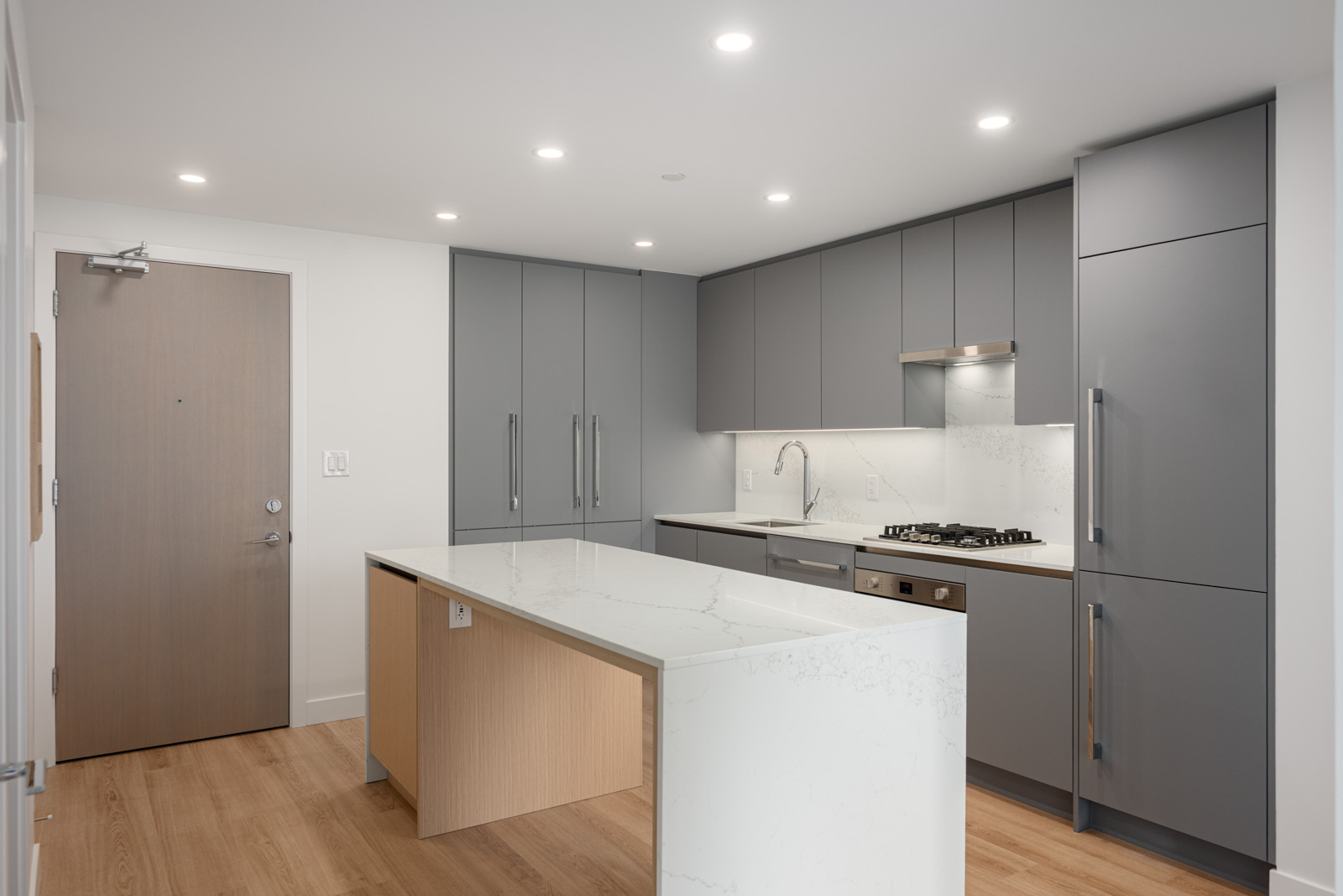 Modern kitchen with gray cabinets, stainless steel appliances, a built-in oven, marble island, and wood flooring, illuminated by recessed ceiling lights.
