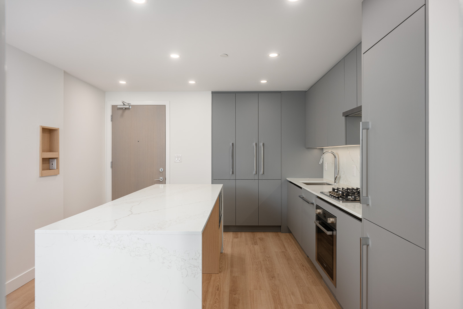 Modern kitchen with gray cabinets, built-in appliances, a marble island countertop, wooden flooring, and recessed ceiling lights.