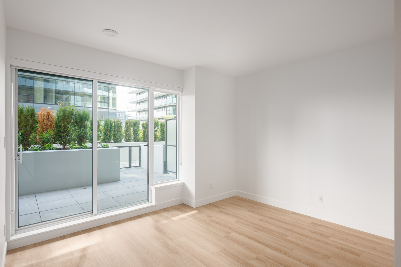 Empty, unfurnished room with light wood flooring, white walls, and large sliding glass doors opening to a patio with greenery and a modern building in the background.