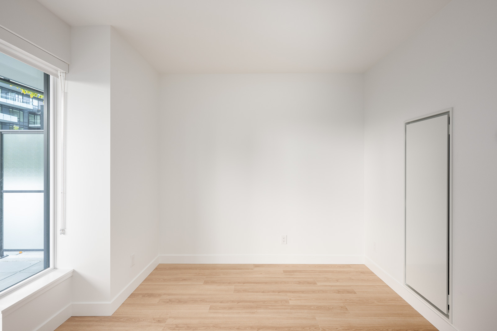 Empty, unfurnished room with white walls, light wood flooring, a large window on the left, and a closed door or access panel on the right wall.