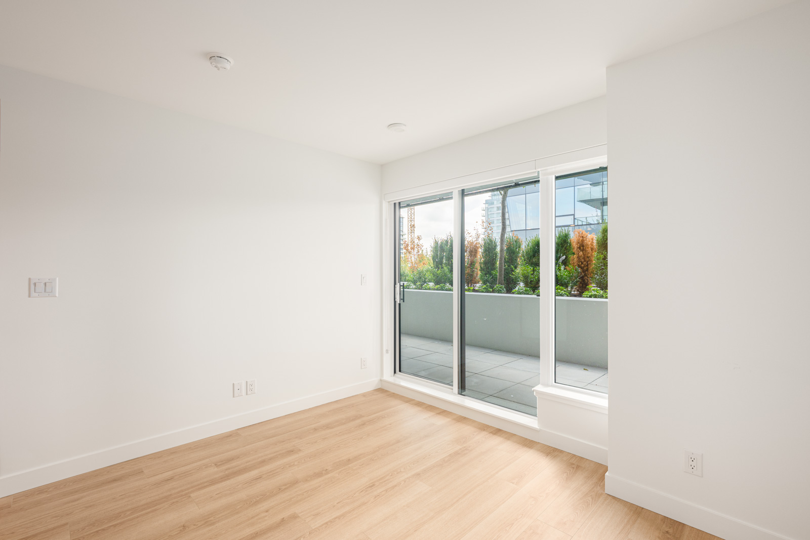 Empty, unfurnished room with light wood flooring, white walls, and large sliding glass doors leading to a balcony with plants outside.