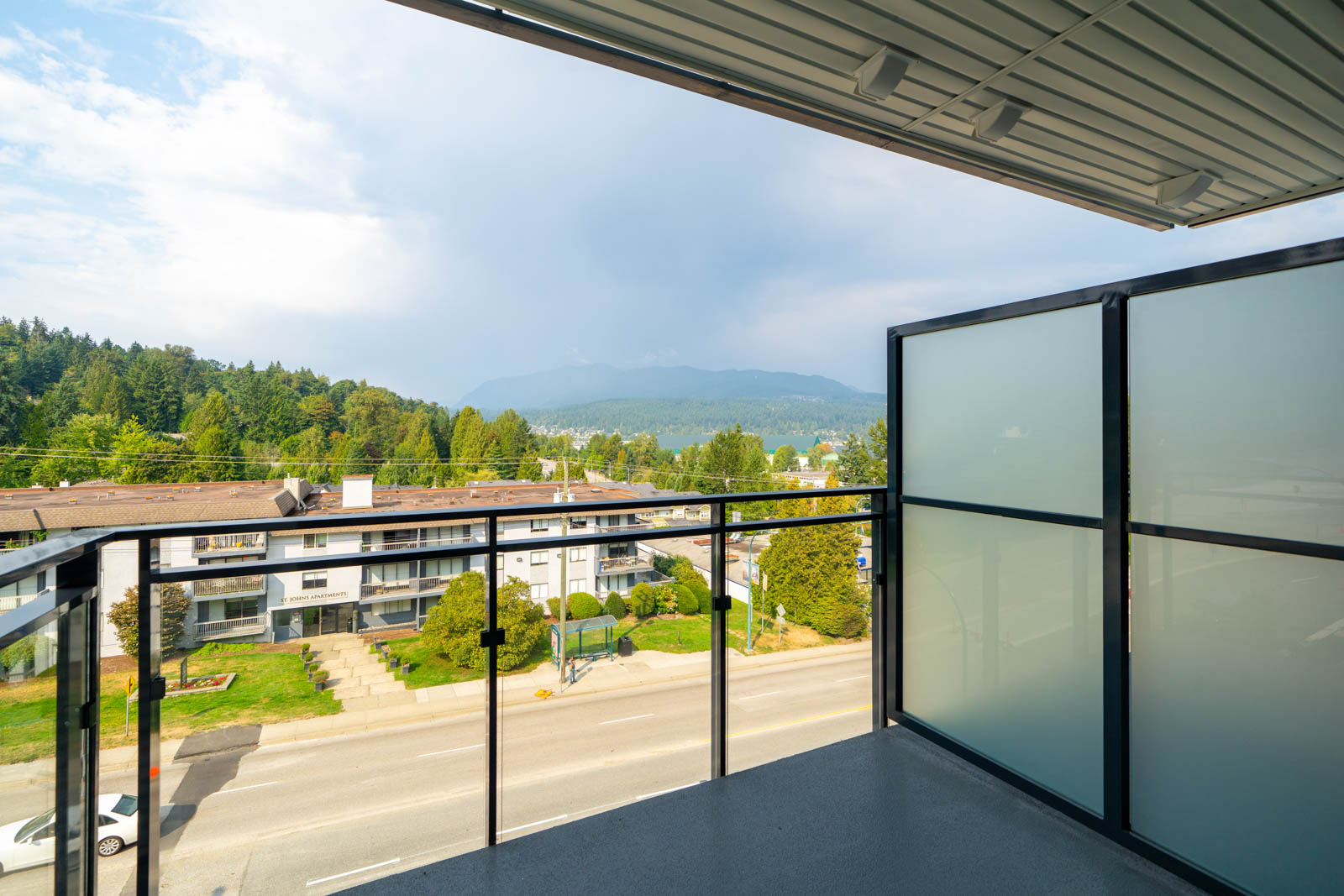 View from a modern balcony with glass railing overlooking a street, residential buildings, trees, and distant mountains under a partly cloudy sky.